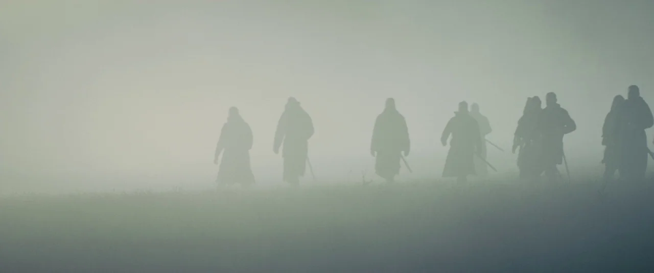 Macbeth (2015) movie still: wide — A group of figures in medieval-style cloaks walk across a grassy field in thick fog. The silhouettes move from…