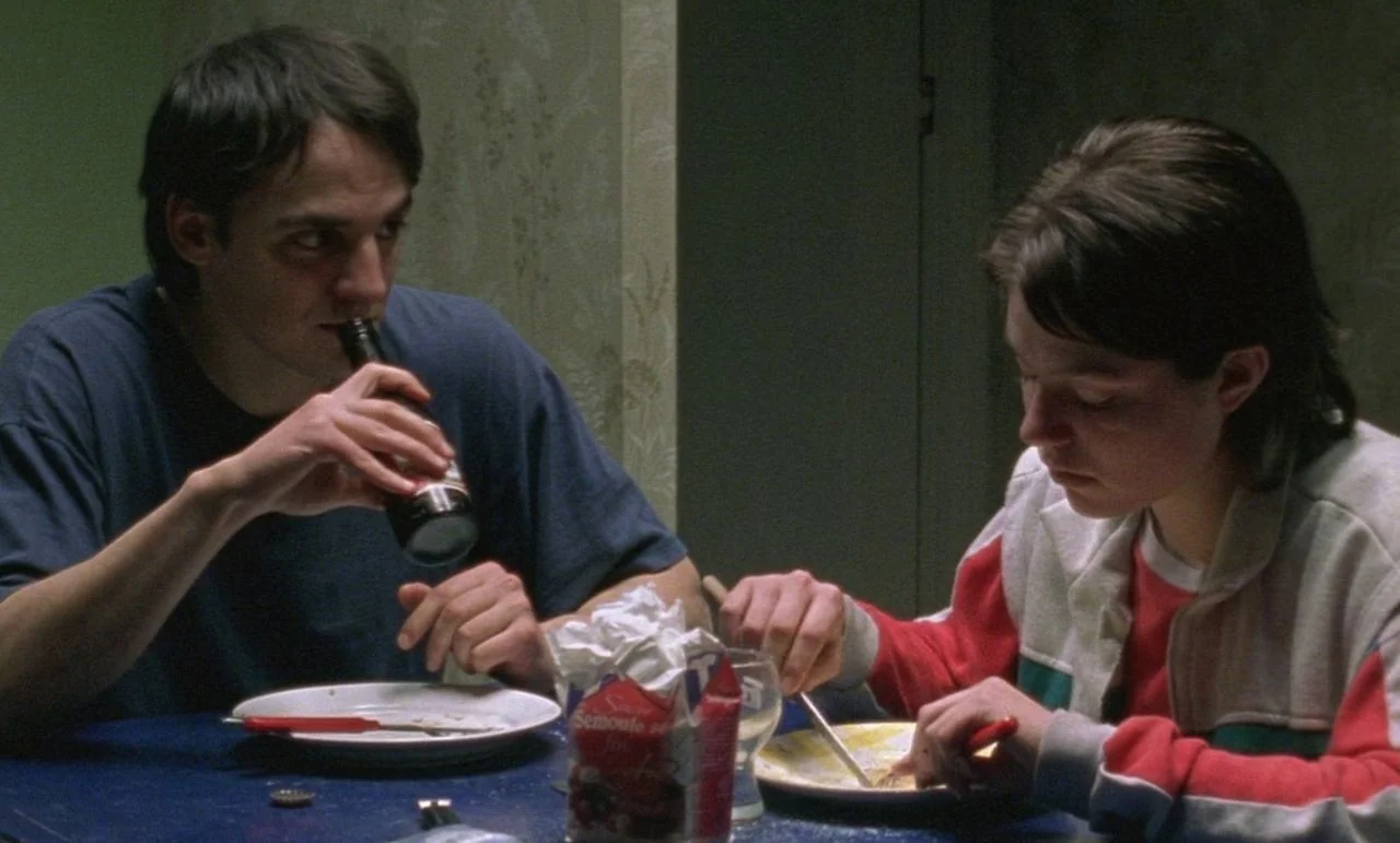 Rosetta (1999) movie still: medium — Two individuals sit at a blue table covered in plates and food items. The man drinks from a bottle while the…