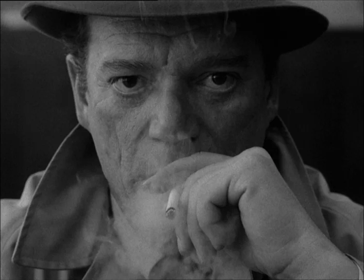 Alphaville (1965) movie still: closeup — A man wearing a hat and coat occupies a dark interior space. He holds a lit cigarette to his lips while…
