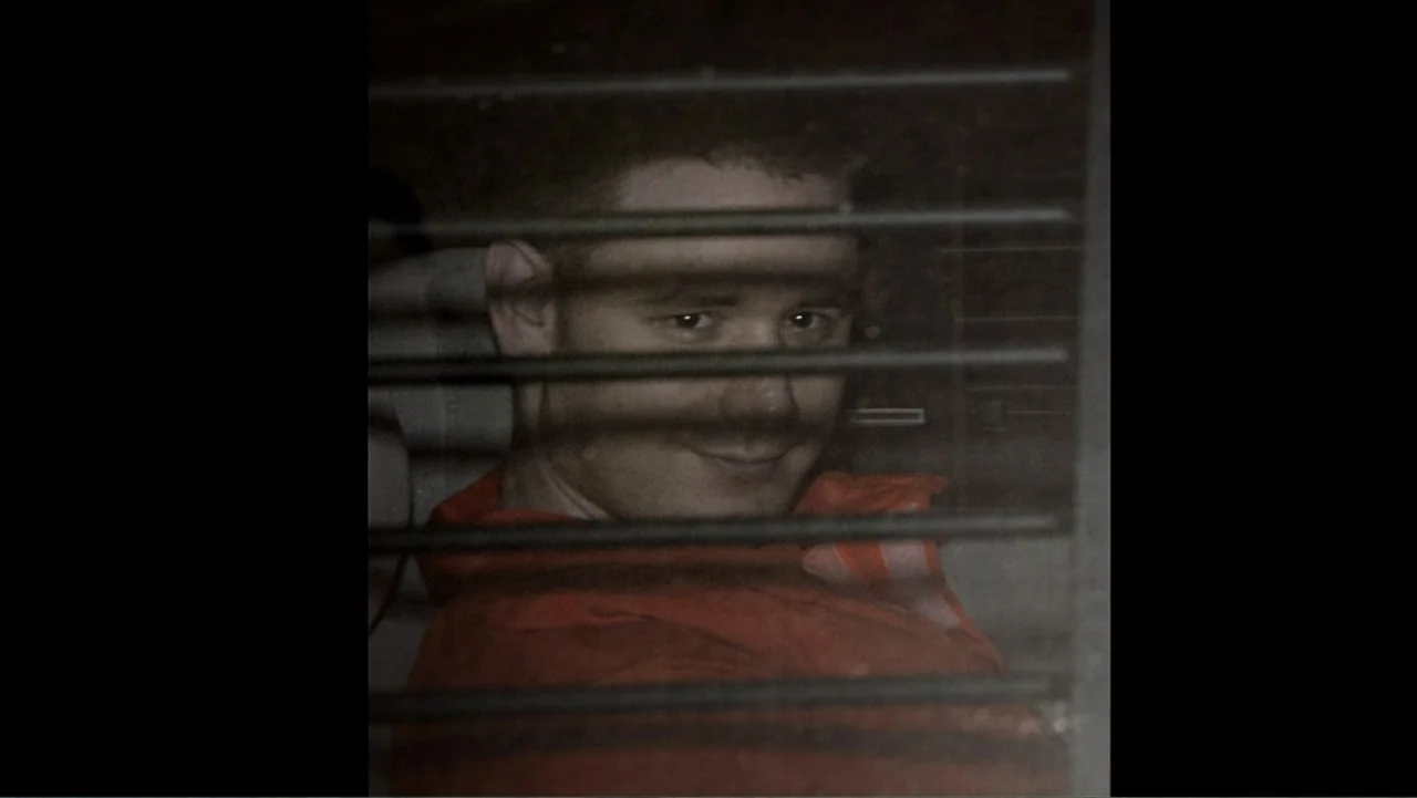 The Cheshire Murders (2013) movie still: closeup — A man looks through horizontal blinds from the interior of a darkened vehicle. The subject wears…