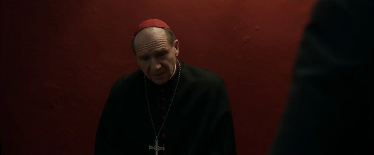 Conclave (2024) movie still: medium — A man in clerical vestments stands before a textured red wall. The cardinal gazes downward in a dark interior…