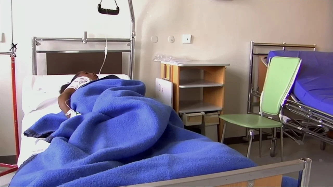 Sister (2014) movie still: medium — A minimalist clinical patient room features a metal bed frame and a plastic chair. A young girl rests under a…