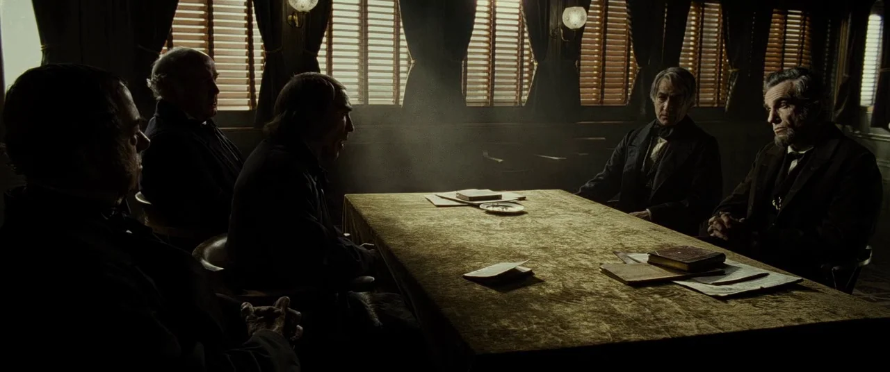 Lincoln (2012) movie still: wide — A dark, wood-paneled room features a long table covered in a worn gold cloth. Five men in formal 19th-century…