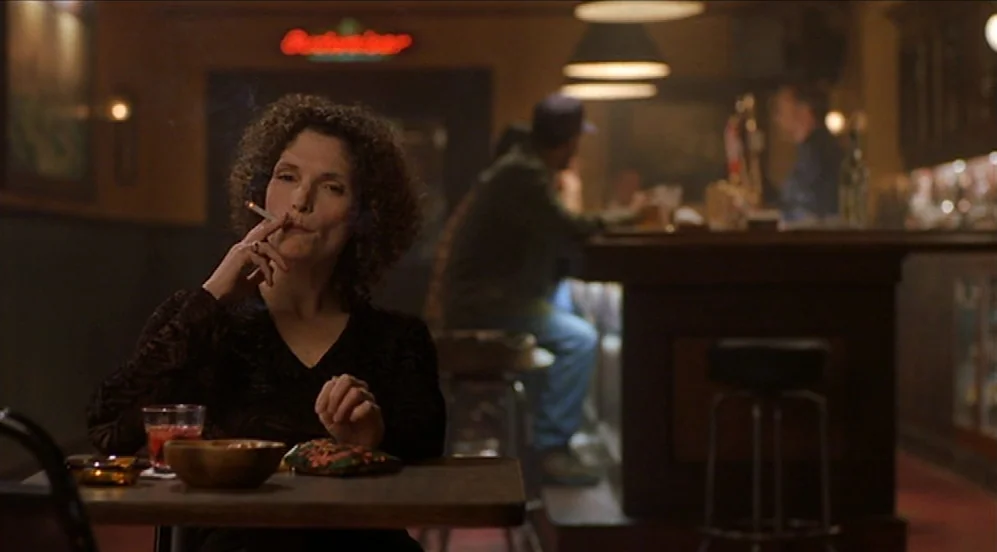Limbo (1999) movie still: medium — A dimly lit tavern with a wooden counter and a glowing Budweiser neon sign. A woman with dark curly hair sits at…