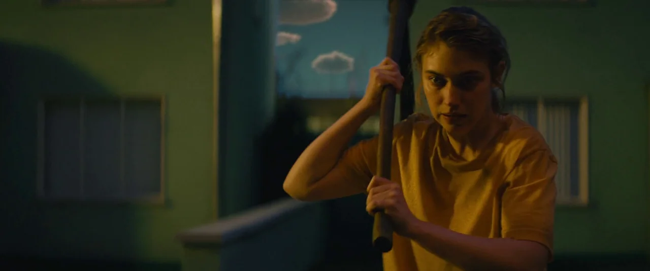 Vivarium (2019) movie still: medium — An apartment building exterior features green walls at night under a darkening sky. A young woman grips a…