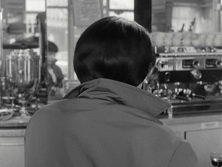 Vivre Sa Vie (1962) movie still: medium — A man sits at a counter in a diner seen from behind. An espresso machine and glass objects are visible on…
