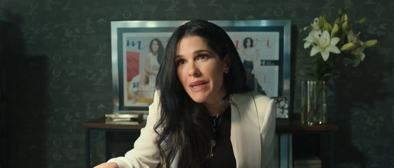 Cindy La Regia (2020) movie still: medium — A professional office space features dark patterned wallpaper and a framed magazine cover background. A…