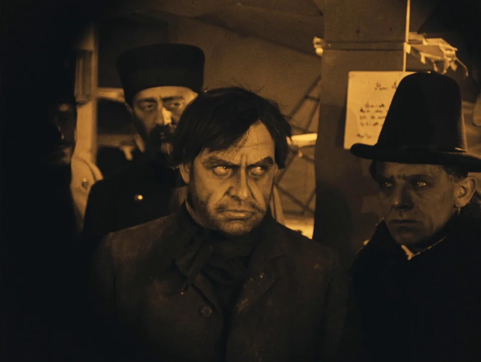 The Cabinet of Dr. Caligari (1920) movie still: medium — A dimly lit, cramped interior space with a wooden post and paper sign background. Three men…