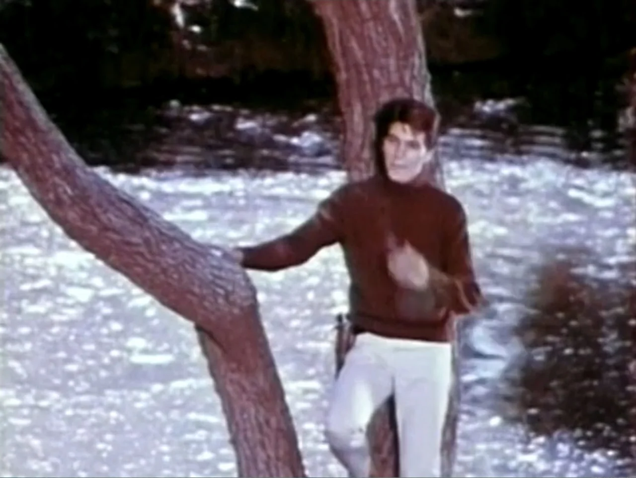 In the Year 2889 (1969) movie still: medium — A man stands outdoors near a brightly lit, flowing river. He leans casually against the rough bark of…