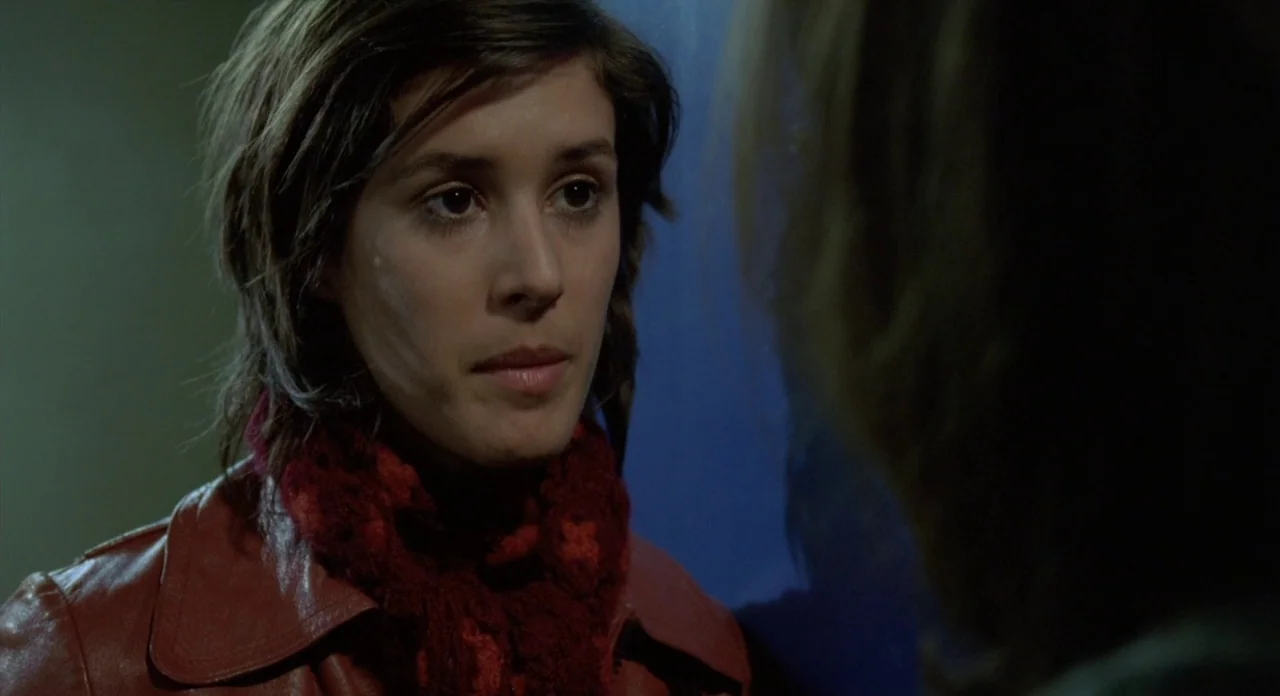 Read My Lips (2001) movie still: closeup — A dimly lit indoor space provides a dark, indistinct backdrop for a two-shot interaction. A young woman…