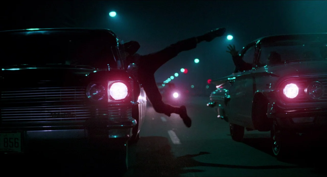 I Wanna Hold Your Hand (1978) movie still: wide — A night scene on an open asphalt road between two vintage cars. A man is mid-air while jumping…