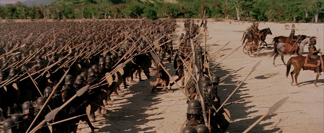 Troy (2004) movie still: wide — A vast ancient army marches in rigid formation across a wide sandy desert plain. Thousands of soldiers in bronze…