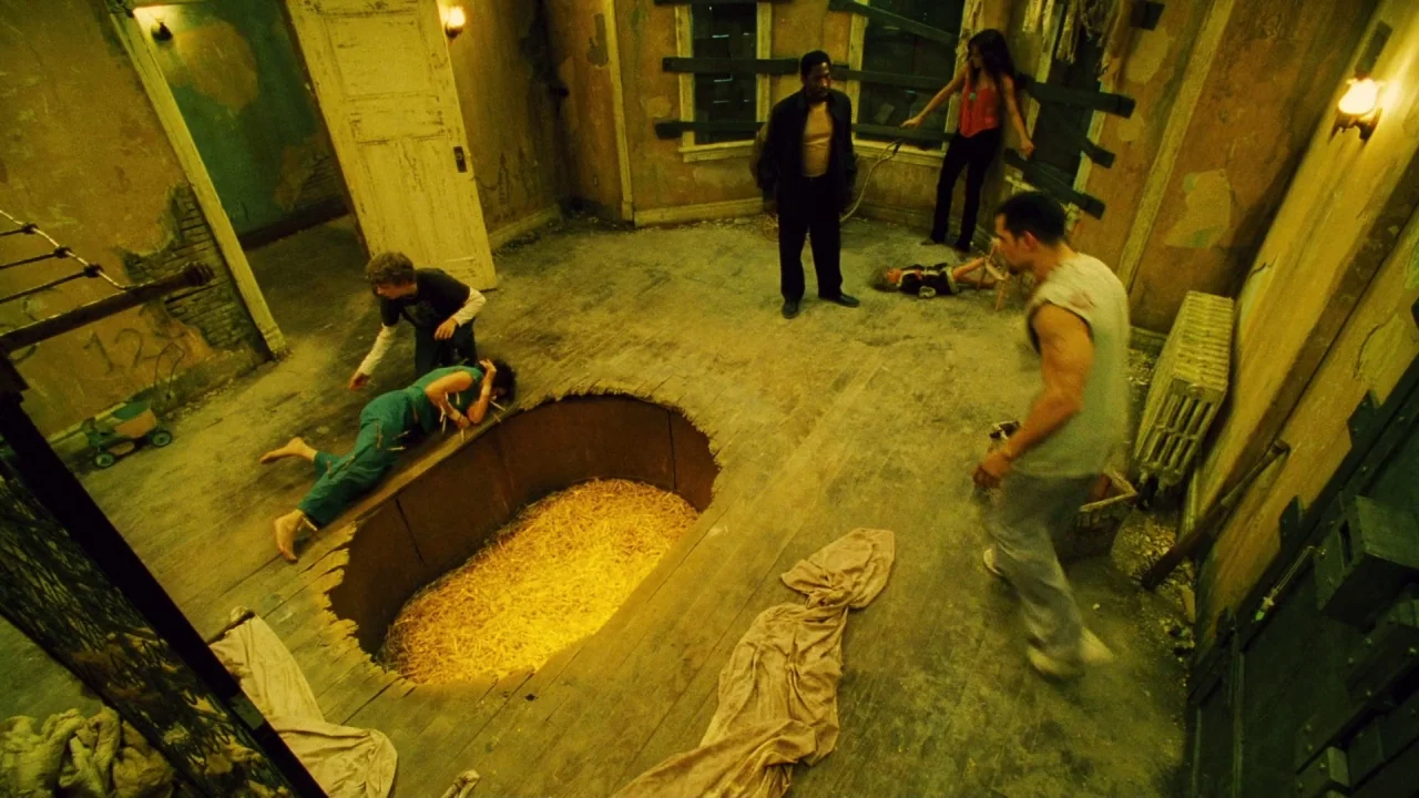 Saw II movie still: wide — A dilapidated, desolate space features peeling paint and boarded-up windows. Multiple individuals surround a large,…