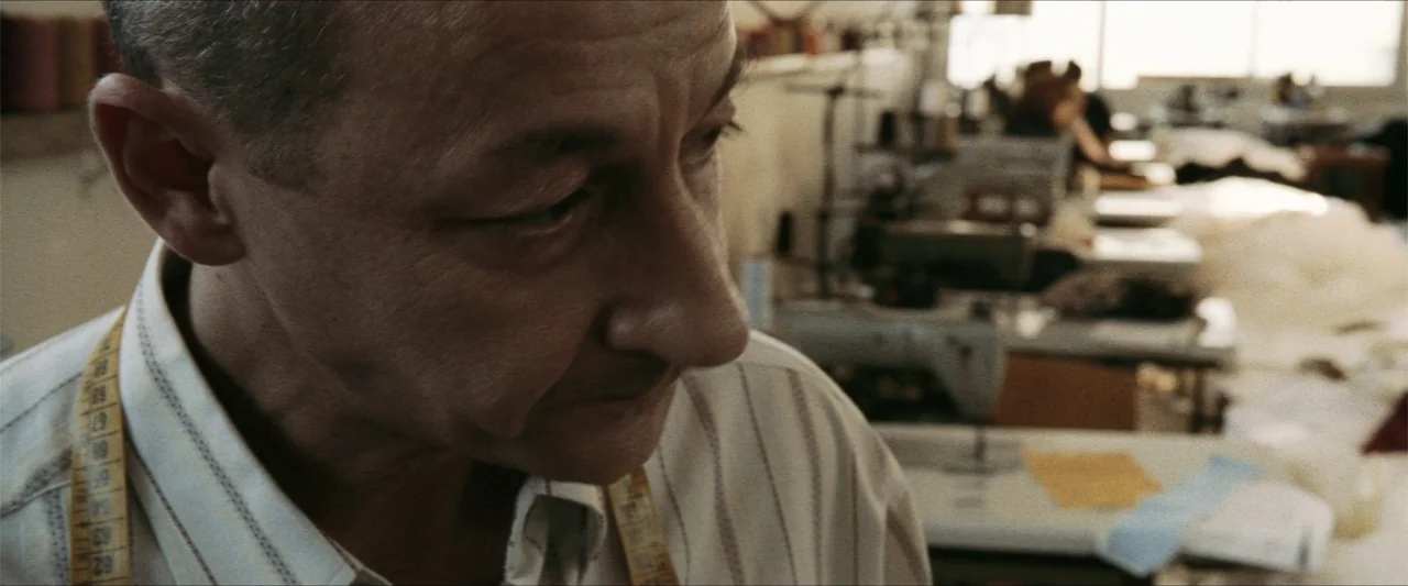 GOMORRAH (2023) movie still: closeup — A sunlit industrial sewing workshop features rows of machinery and white fabric stacks in background. An…