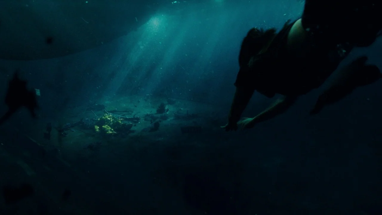 Bumblebee (2018) movie still: wide — Underwater scene showing the dark seafloor and debris under a sunlit surface. A swimmer approaches a bright…