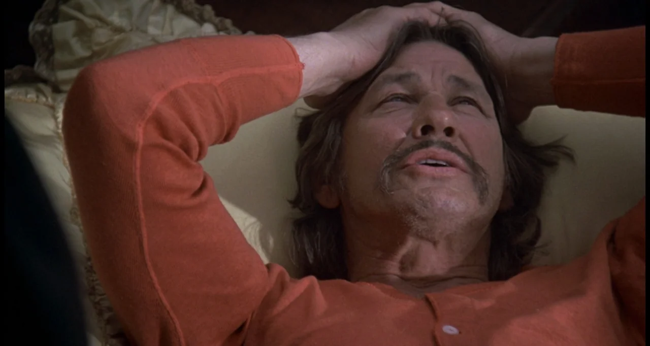 The White Buffalo (1977) movie still: closeup — A man rests on a cream-colored pillow inside a bedroom setting. He is wearing an orange long-sleeved…