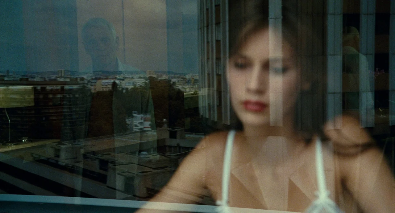 Young & Beautiful (2013) movie still: medium — A woman sits in a room with a large pane of glass overlooking a city. Her reflection is cast upon the…