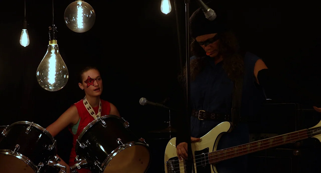 Hellbender (2021) movie still: medium — A dark, dimly lit stage features two musicians against a black backdrop. Hanging exposed filament light…