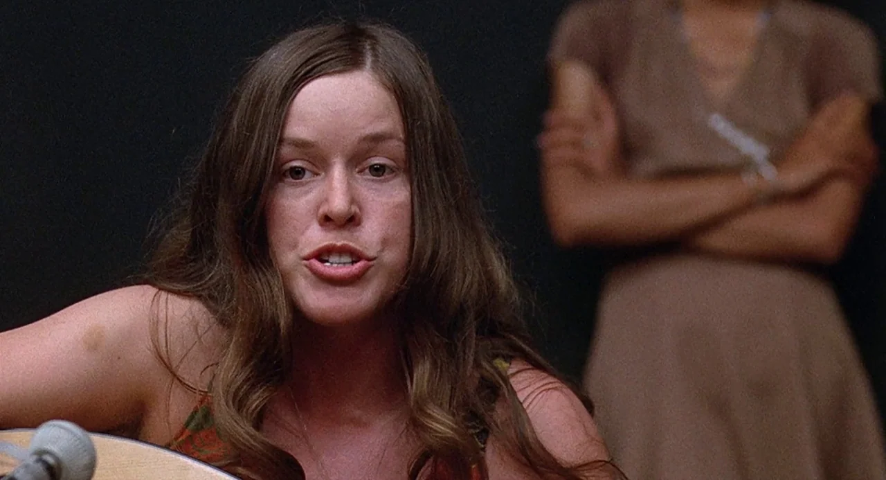 Taking Off (1971) movie still: closeup — A dimly lit room serves as a recording space with dark walls. A young woman sings into a microphone while…