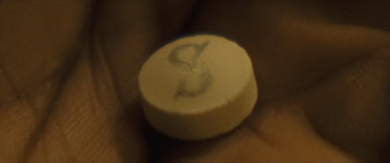 Synchronic (2019) movie still: closeup — A small, pale circular pill with a stamped letter sits in a person's palm. The surface of the skin is…