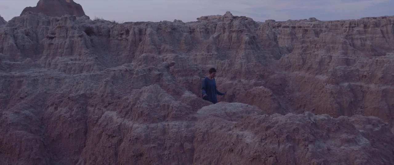 Nomadland (2020) movie still: wide — A vast, eroded landscape of sedimentary rock formations under a twilight sky. A person walks through the uneven…