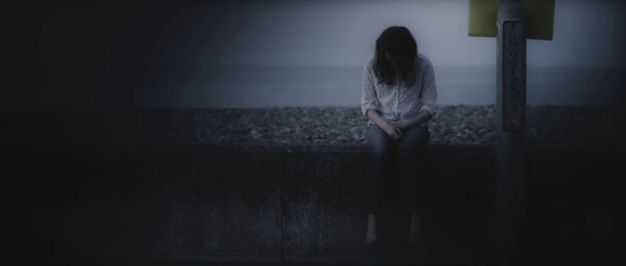 A Dark Song (2016) movie still: fullbody — A dark concrete seawall runs horizontally across the frame at the water's edge. A woman sits on the wall…