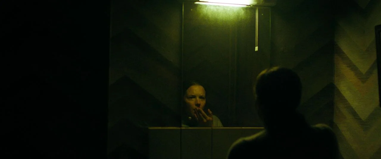 Saint Maud (2019) movie still: medium — A dimly lit room features a chevron-patterned wallpaper behind a central rectangular wall mirror. A person…