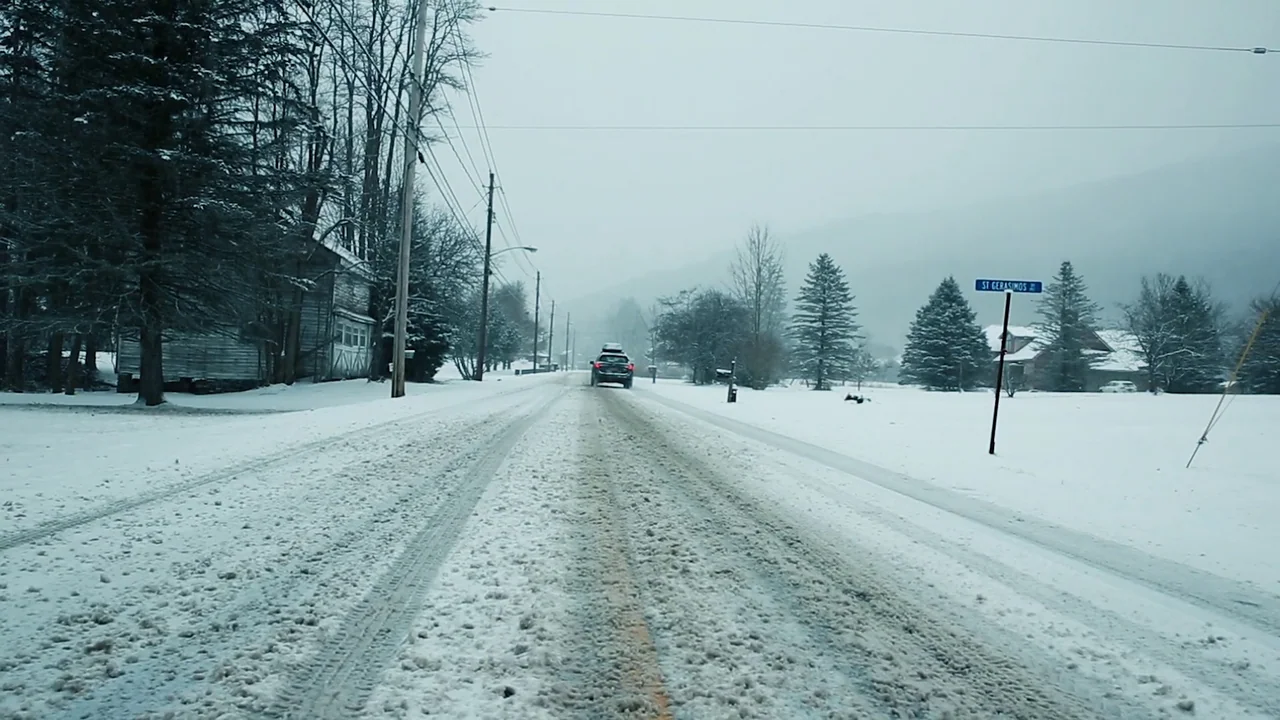 The Deeper You Dig (2019) movie still: wide — A rural road covered in packed snow leads toward trees under an overcast sky. A dark SUV with a roof…