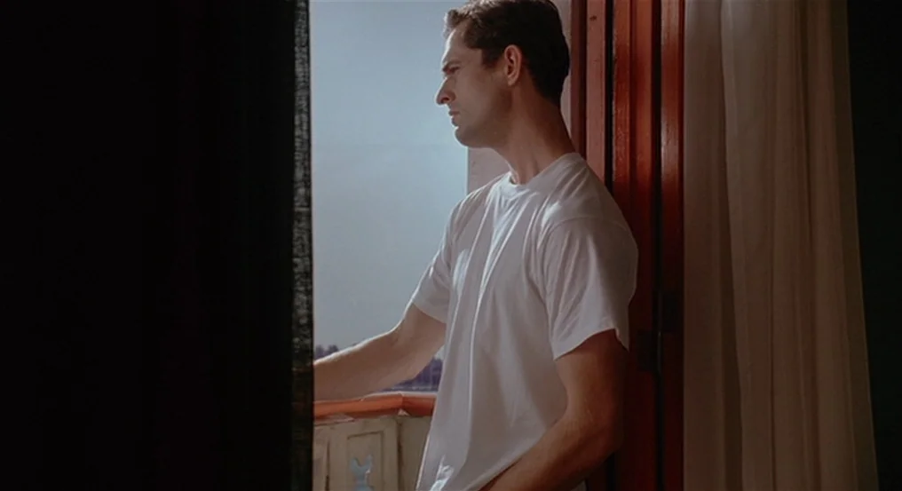 The Comfort of Strangers (1990) movie still: medium — A man stands inside a room next to an open glass balcony door. He faces the bright exterior…