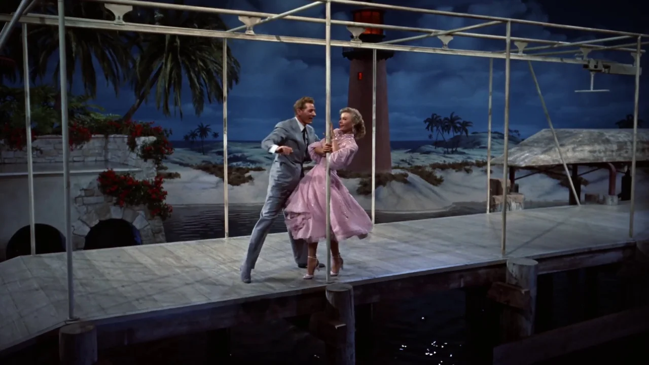 White Christmas (1954) movie still: fullbody — A painted theatrical backdrop depicts a coastal scene with a lighthouse and palm trees. A man in a…