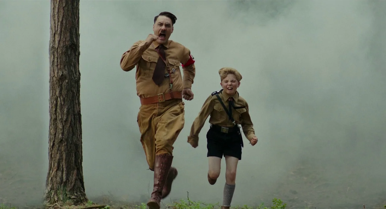 Jojo Rabbit (2019) movie still: fullbody — A man and a boy in tan military uniforms run together through a wood. The air behind them is filled with…