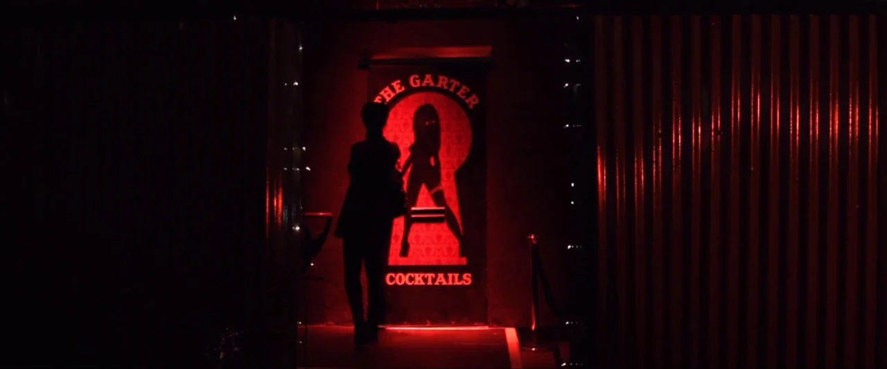 Girl Walks Into a Bar (2011) movie still: wide — A dark outdoor space features a glowing red doorway displaying a keyhole logo. A woman walks toward…