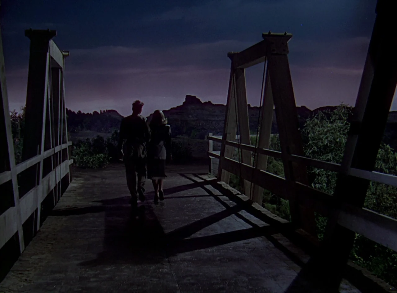 Desert Fury (1947) movie still: wide — A wooden truss bridge crosses over dark terrain under a twilight night sky. A man and a woman walk away from…