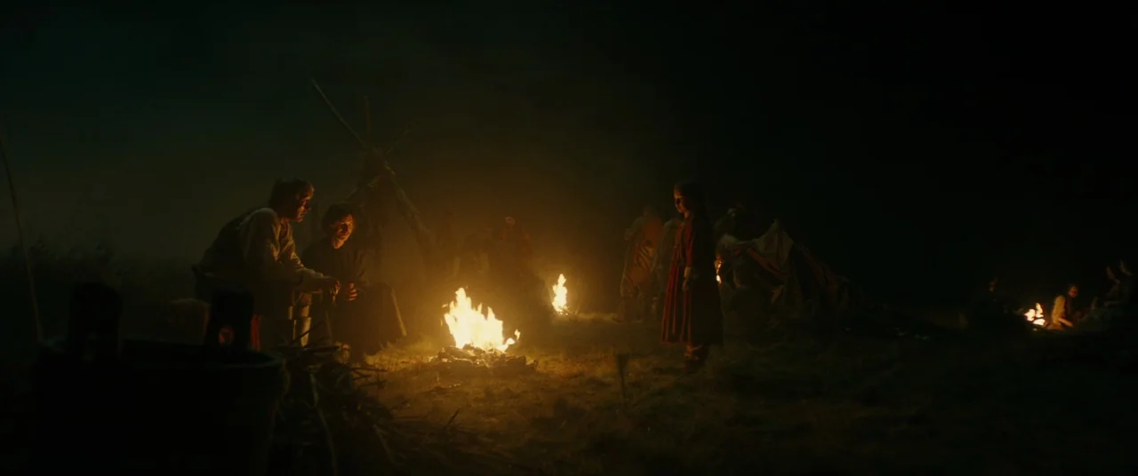 The Promised Land (2023) movie still: wide — A nighttime encampment features multiple burning fires set in a dark, open field. A child stands…