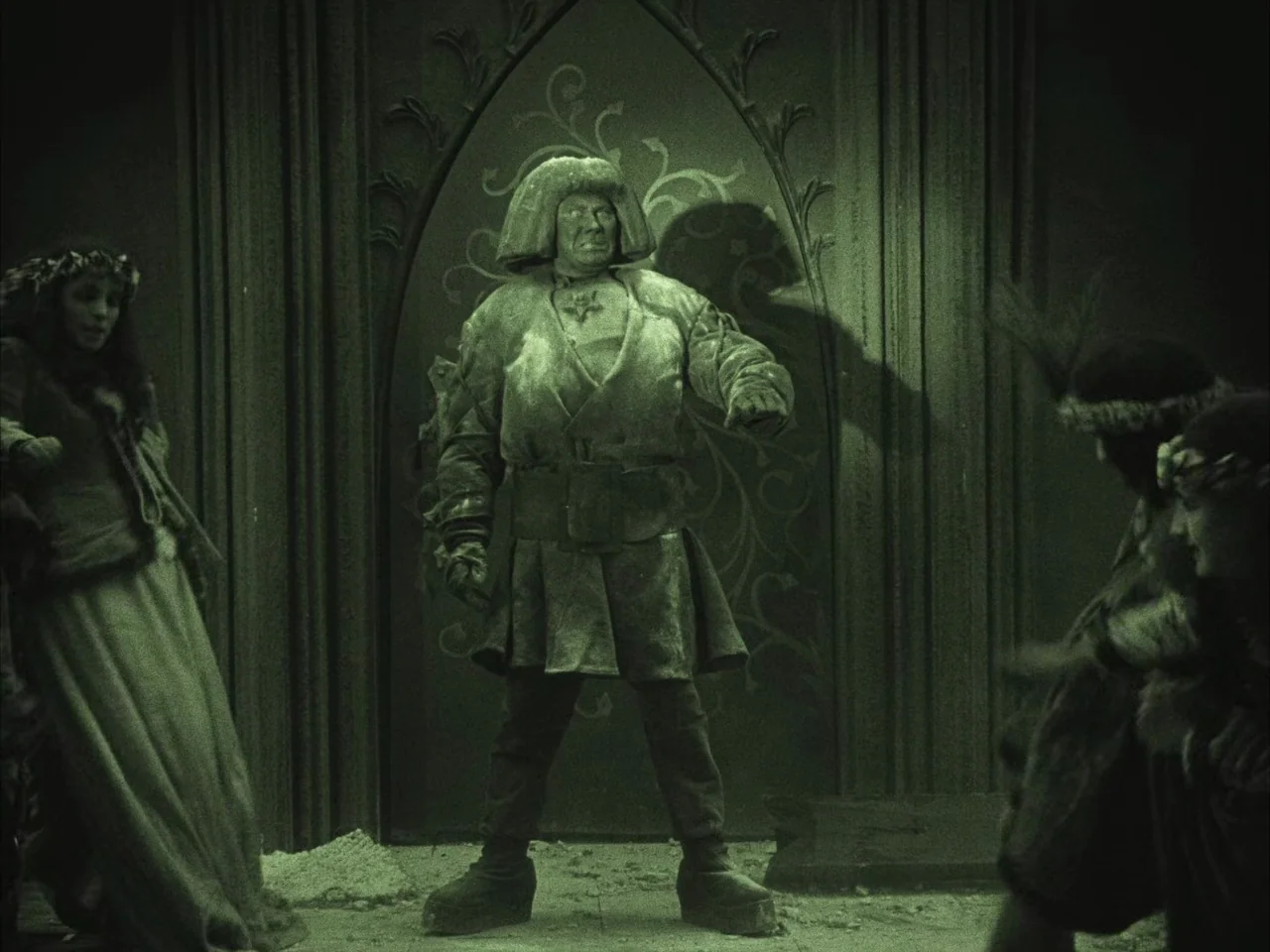 The Golem (1920) movie still: fullbody — A stone chamber features a large ornate archway with decorative metal grillwork. A stocky man wearing a…