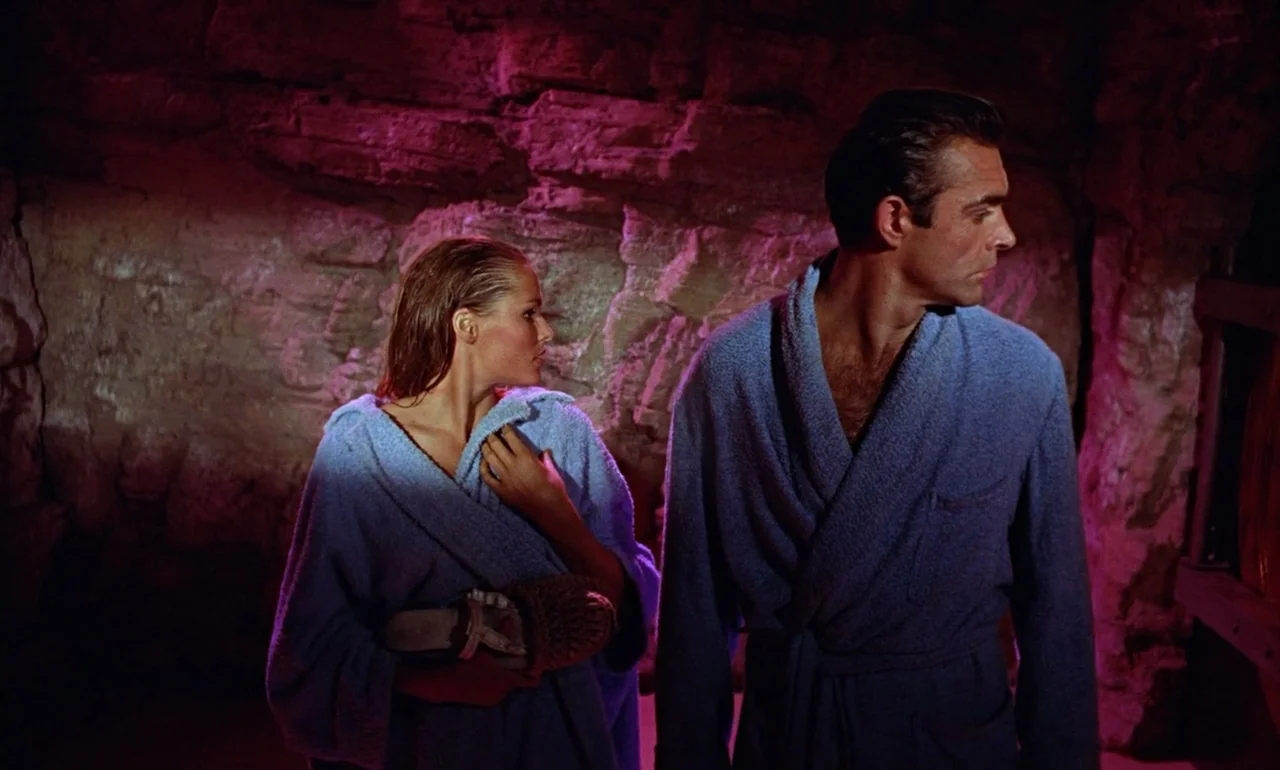 Dr. No (1962) movie still: medium — A dark rocky cavern is illuminated by intense magenta light reflecting off the stone wall. A man and a woman…