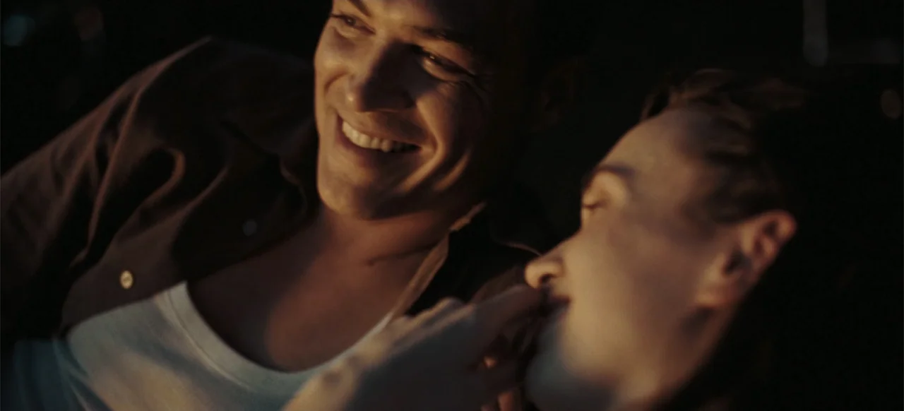 The Tank (2025) movie still: closeup — Two men lie together in a darkened room with very little visible surrounding detail. One man looks at the…