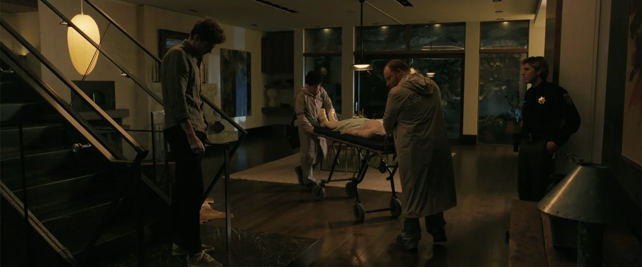 Companion (2025) movie still: wide — A spacious modern home interior with dark hardwood floors and large glass walls. Two medics transport a person…