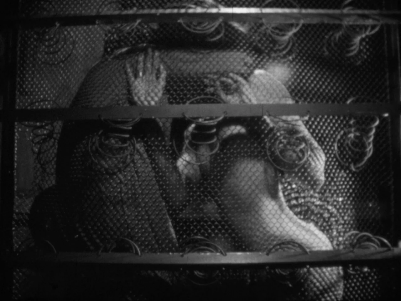 A Woman Kills (1968) movie still: medium — A dark bedroom viewed through the metal coils of an exposed mattress spring. Two individuals embrace…