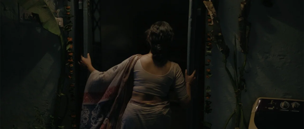 Nishaanchi 2 (2025) movie still: medium — A dimly lit doorway opens onto a dark exterior at night. A woman in a traditional sari pushes the door…
