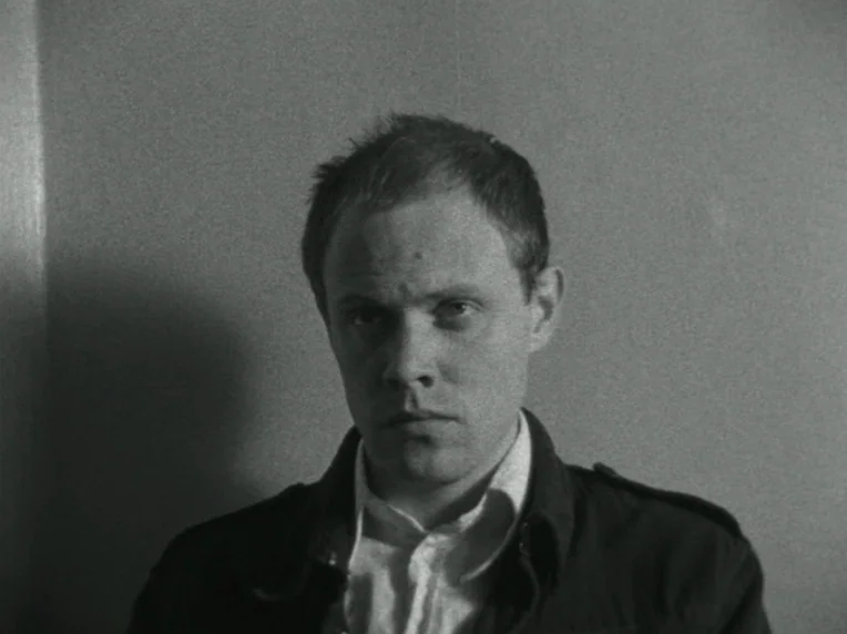 Children (1976) movie still: closeup — A man stands centered against a flat, featureless wall in an indoor space. He stares blankly at the camera…