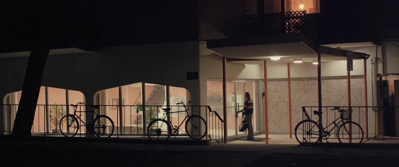 The Editor (2014) movie still: wide — A dark night exterior shows the glass entrance of an apartment building with metal railings. A man standing…