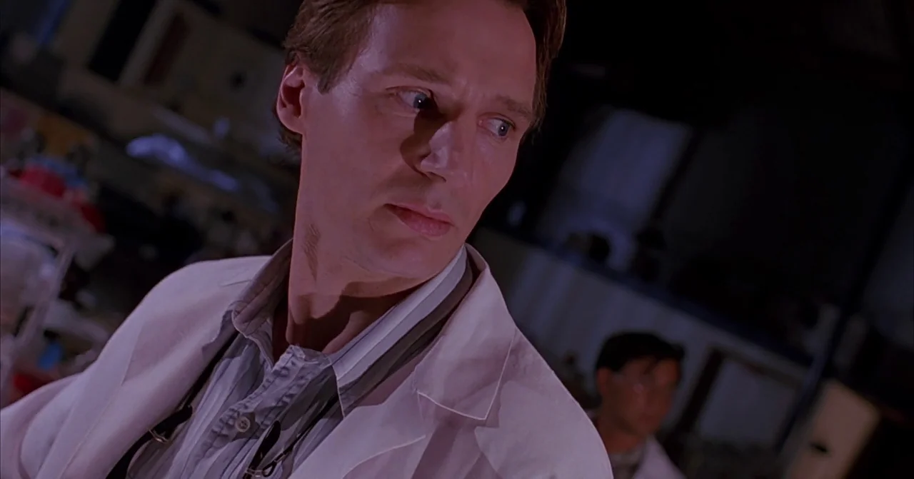 Darkman (1990) movie still: closeup — A dimly lit room with laboratory equipment visible in the blurred background. A man in a white lab coat looks…