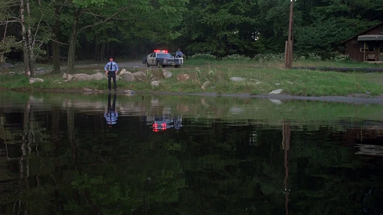 Friday the 13th (1980) movie still: wide — A dark lake reflects the trees and a police car parked on the bank. An officer in uniform stands at the…