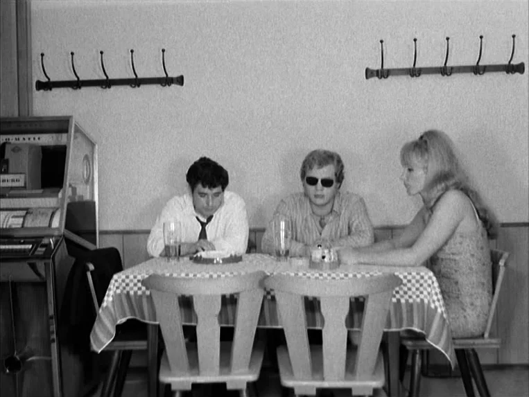 Katzelmacher (1969) movie still: medium — A modest eatery with a checkered tablecloth and wooden chairs set against a blank wall. Two men and one…