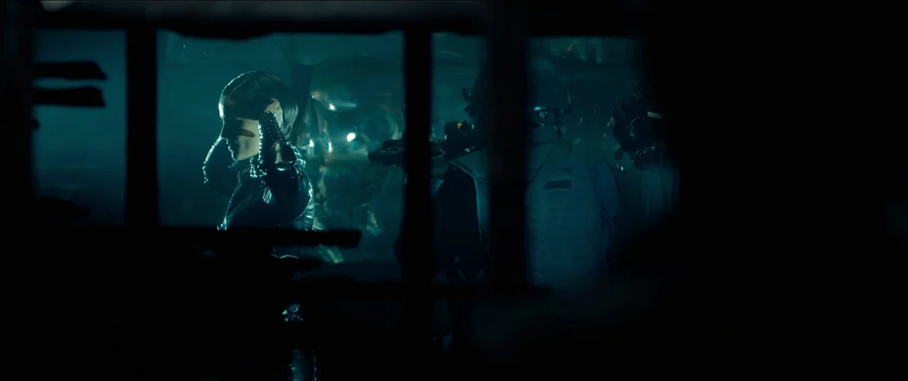 The Matrix Resurrections (2021) movie still: medium — An industrial space with dark framing elements separating the interior view. A woman in black…