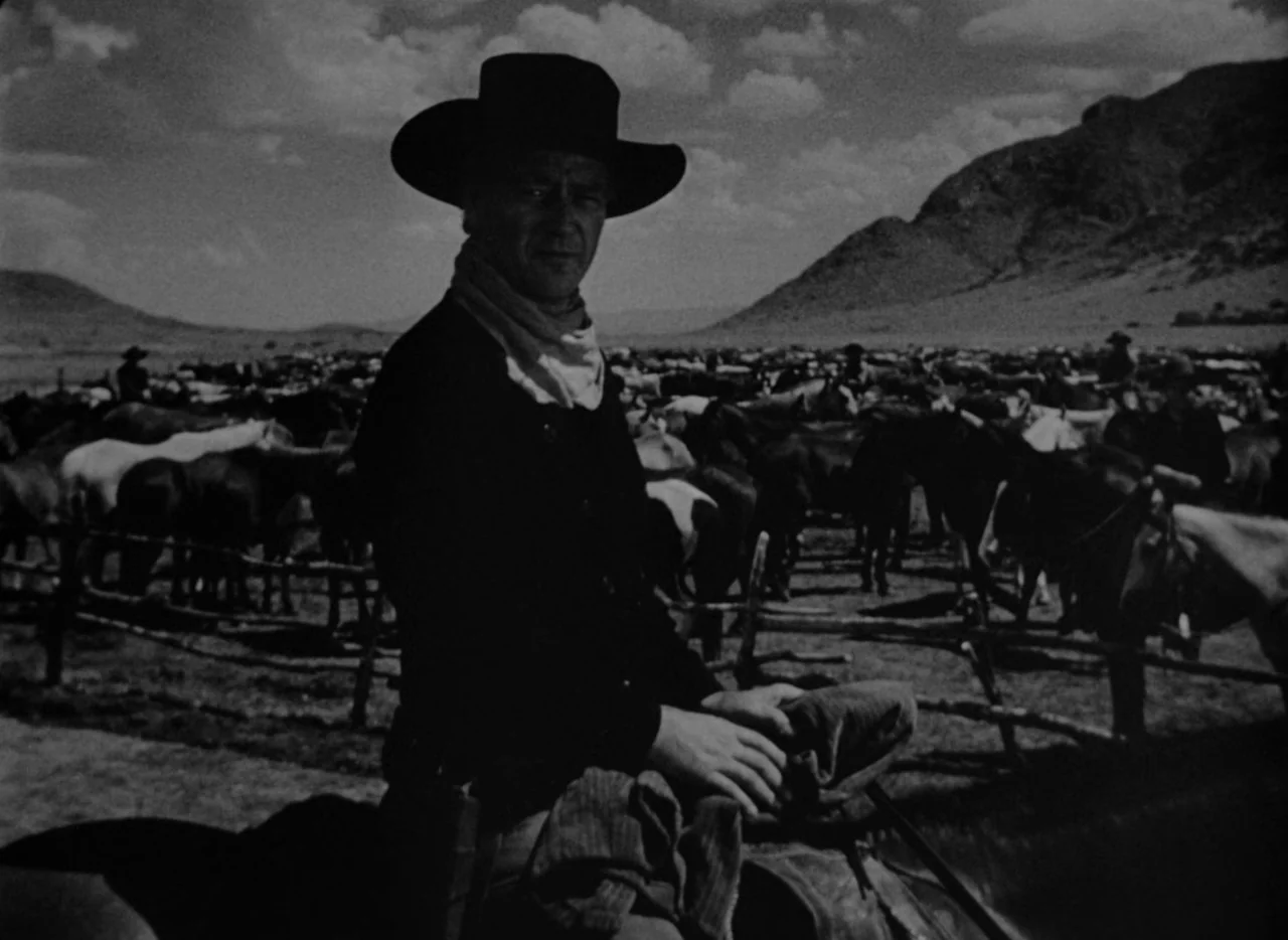 Red River (1948) movie still: medium — A vast, sunlit horse corral with a rugged mountain landscape in the distance. A man in a cowboy hat sits on…