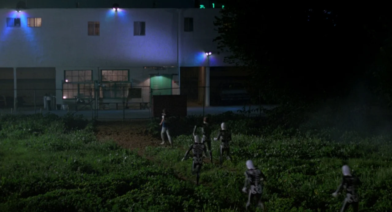 The Karate Kid (1984) movie still: wide — An open grassy field at night sits in front of a white multi-story building. Several figures wearing…