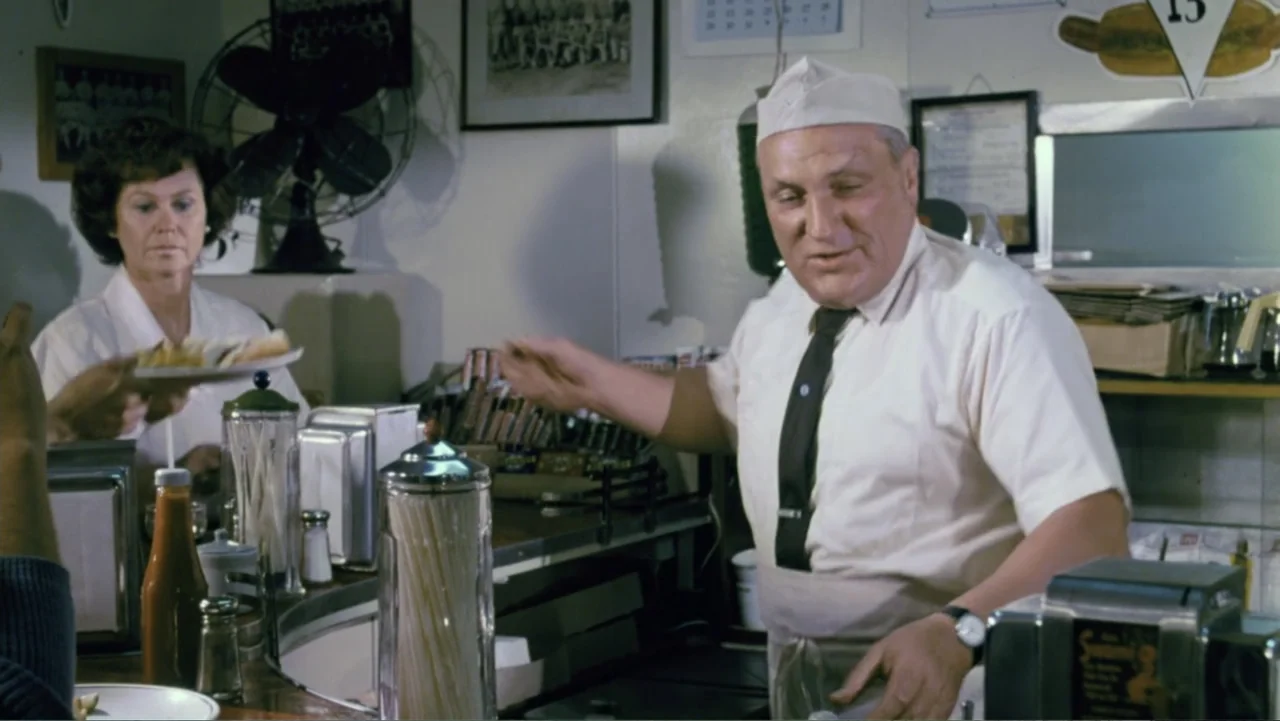 Aunt Mary (1979) movie still: medium — A classic mid-century diner interior features a white counter, metal napkin dispensers, and framed photos. A…