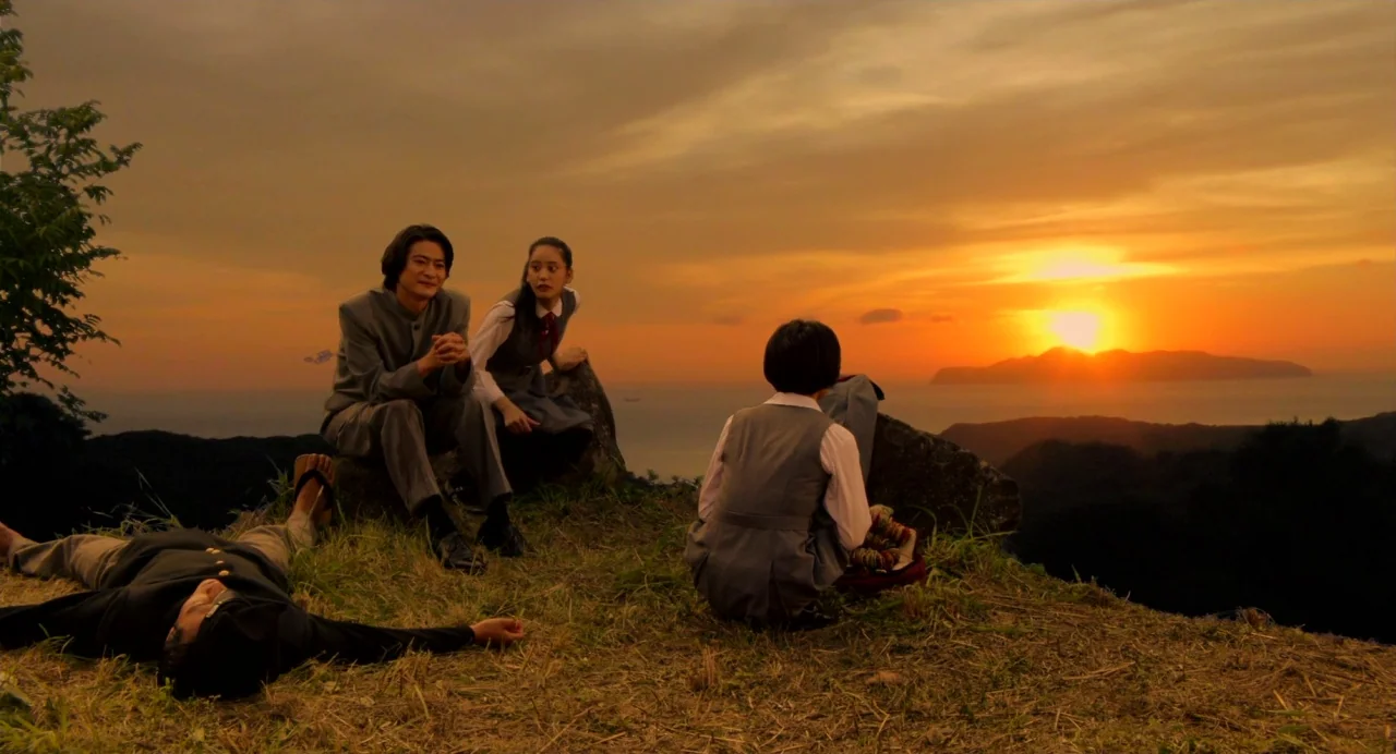 Hanagatami movie still: wide — A grassy hill overlooks a wide bay with distant mountains during a vibrant sunset. Four students rest on the terrain,…